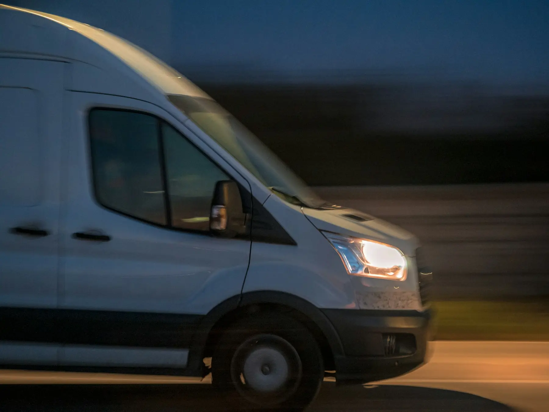 A white removal van driving at night with its headlights on, blurred by speed
