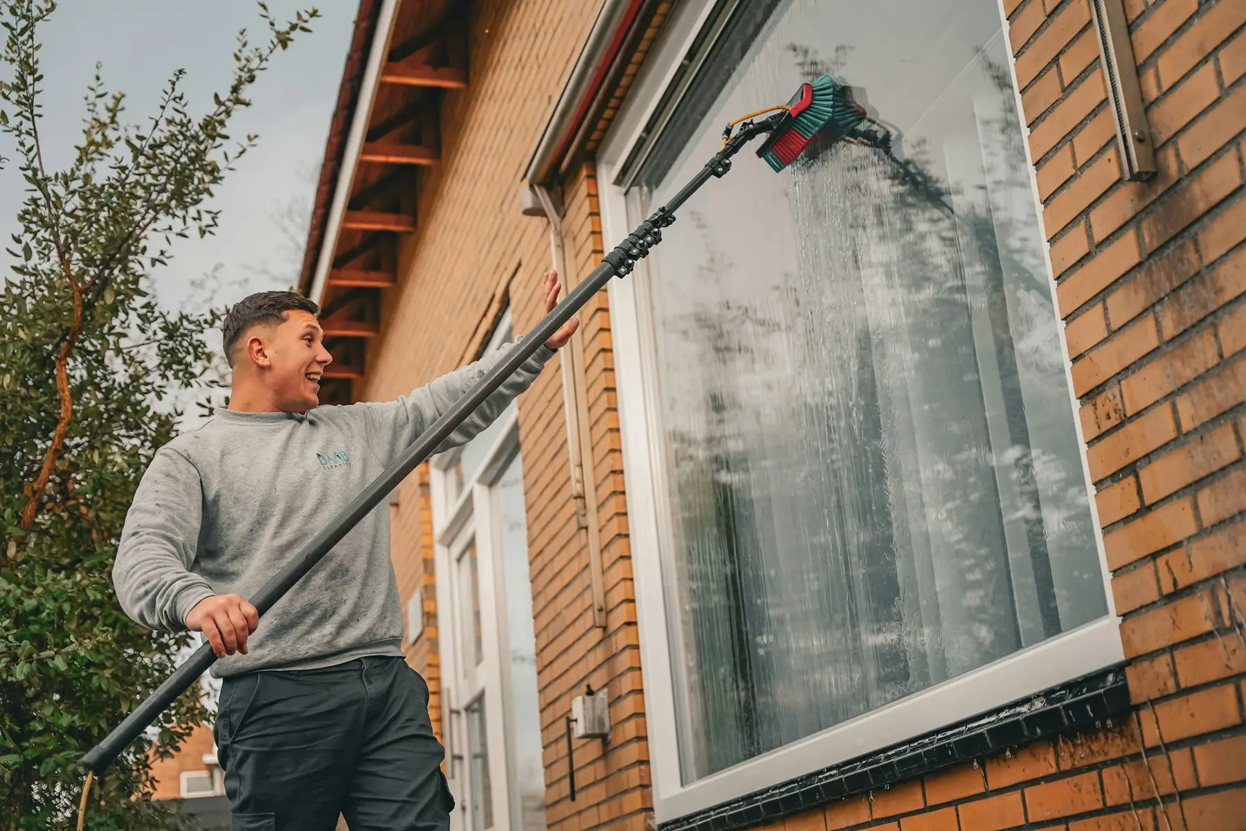 A smiling window cleaner washing a first-floor window with a water-fed pole