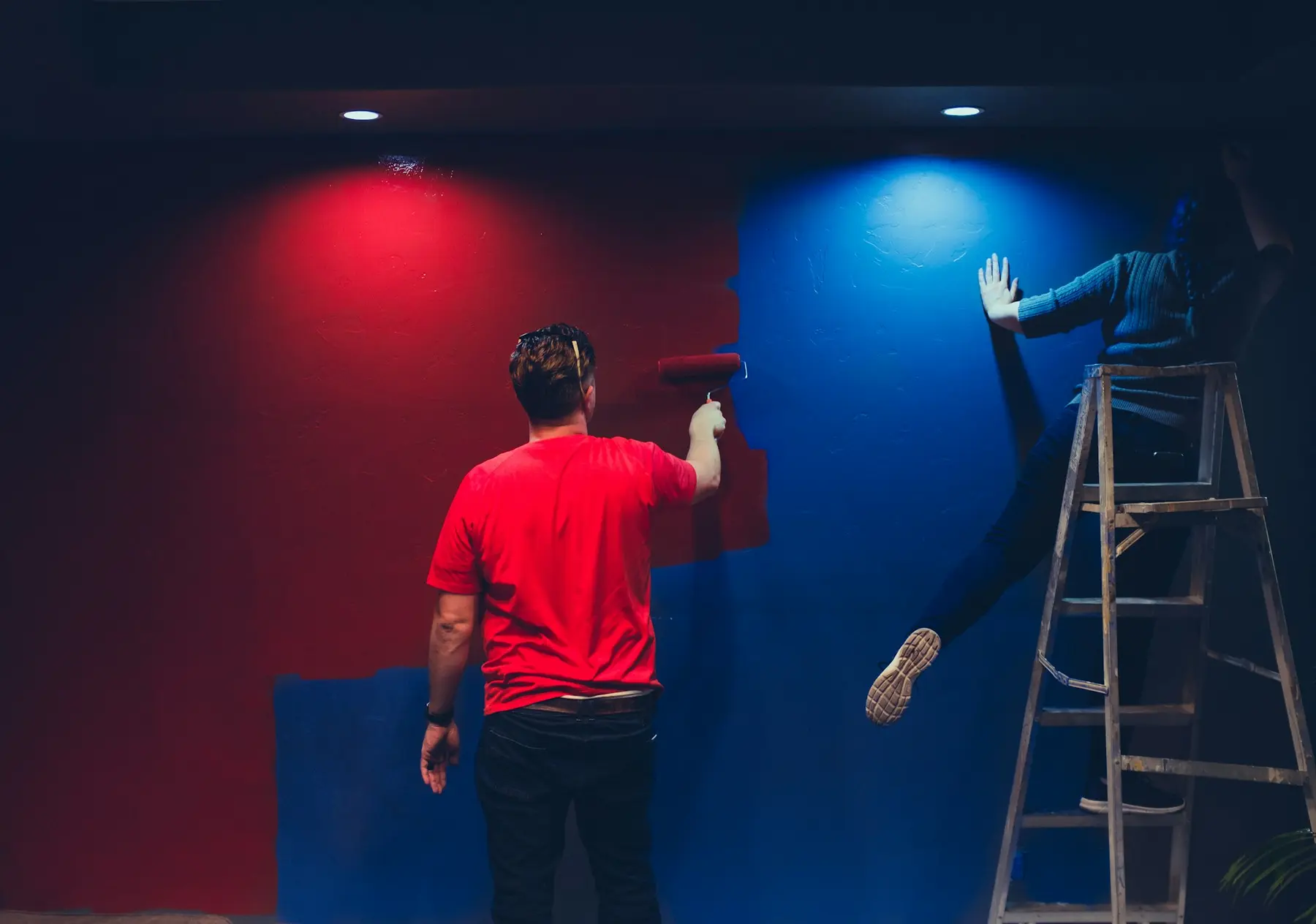 A decorator in a red T-shirt painting a wall red next to a blue wall and a stepladder